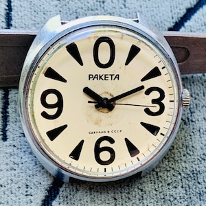 Puede incluir: Un reloj de pulsera Raketa de tono plateado con esfera color crema, que presenta grandes números negros en las posiciones 0, 3, 6 y 9, manecillas negras y el texto "PAKETA" y "СДЕЛАНО В СССР" impreso en negro en la esfera.