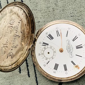 Puede incluir: Reloj de bolsillo antiguo abierto con caja plateada y esfera blanca. La esfera presenta números romanos negros, un pequeño dial de segundos y el texto "Георги Фавра Жанко". La parte trasera abierta del reloj tiene texto grabado.