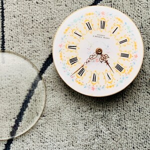 K&ouml;nnte beinhalten: Ein antikes Taschenuhrzifferblatt mit r&ouml;mischen Ziffern, floralen Akzenten und dem Text "MARQUE DE FABRIQUE". Eine klare Glaslinse und ein goldfarbener Uhrenschl&uuml;ssel sind ebenfalls vorhanden.