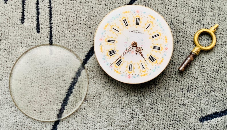 K&ouml;nnte beinhalten: Ein antikes Taschenuhrzifferblatt mit r&ouml;mischen Ziffern, floralen Akzenten und dem Text "MARQUE DE FABRIQUE". Eine klare Glaslinse und ein goldfarbener Uhrenschl&uuml;ssel sind ebenfalls vorhanden.
