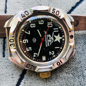 May include: A vintage wristwatch with a black face, silver bezel, and a brown leather strap. The watch face features white numerals, a white star, and the text "Командирские". The watch has a gold-colored crown and a red second hand.