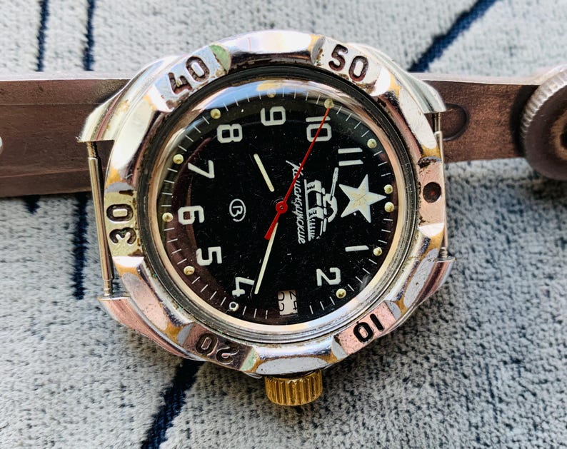 May include: A vintage wristwatch with a black face, silver bezel, and a brown leather strap. The watch face features white numerals, a white star, and the text "Командирские". The watch has a gold-colored crown and a red second hand.