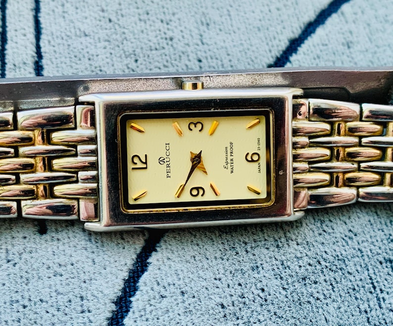 May include: A rectangular Perucci watch with a gold-tone face and hands, featuring gold hour markers and the numbers 12, 3, 6, and 9. The watch has a silver and gold-tone link band. The watch face reads "Expression Water Proof" and "Japan".