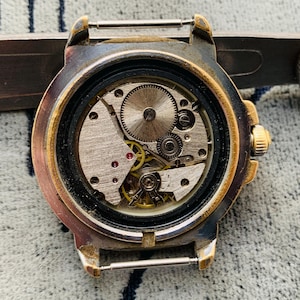 Op de afbeelding: Close-up van een vintage horloge met de achterkant verwijderd, waardoor het ingewikkelde mechanische uurwerk zichtbaar wordt. Het horloge heeft een goudkleurige bezel en een zilverkleurige kast. De interne tandwielen, veren en juwelen zijn zichtbaar.