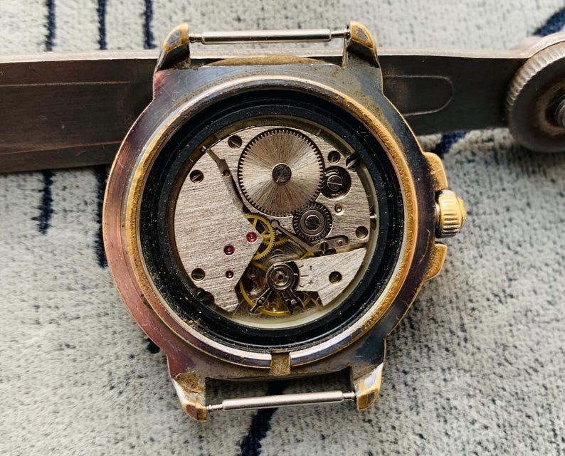 Op de afbeelding: Close-up van een vintage horloge met de achterkant verwijderd, waardoor het ingewikkelde mechanische uurwerk zichtbaar wordt. Het horloge heeft een goudkleurige bezel en een zilverkleurige kast. De interne tandwielen, veren en juwelen zijn zichtbaar.