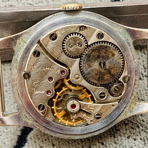 Puede incluir: Primer plano del movimiento de un reloj vintage, mostrando intrincados engranajes, resortes y joyas. Los componentes met&aacute;licos plateados son detallados, con marcas y n&uacute;meros visibles. El mecanismo interno del reloj est&aacute; expuesto.