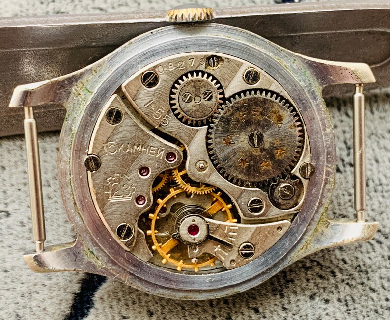 Puede incluir: Primer plano del movimiento de un reloj vintage, mostrando intrincados engranajes, resortes y joyas. Los componentes met&aacute;licos plateados son detallados, con marcas y n&uacute;meros visibles. El mecanismo interno del reloj est&aacute; expuesto.