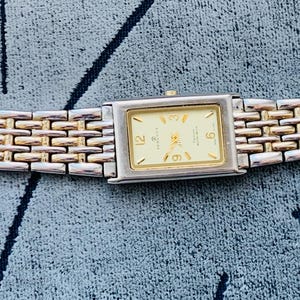 May include: A rectangular wristwatch with a gold-toned face and silver and gold-toned band. The watch face displays the numbers 12, 3, 6, and 9. The watch band is approximately 7 inches long.