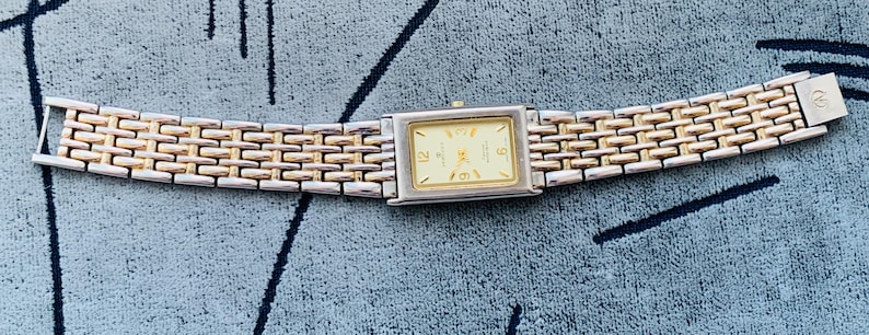 May include: A rectangular wristwatch with a gold-toned face and silver and gold-toned band. The watch face displays the numbers 12, 3, 6, and 9. The watch band is approximately 7 inches long.