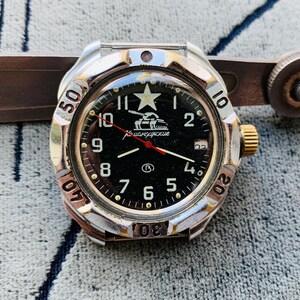 May include: A silver and gold-toned wristwatch with a black face, featuring white numerals and a star emblem. The watch has a red second hand and is set against a textured background. A metal tool is partially visible.