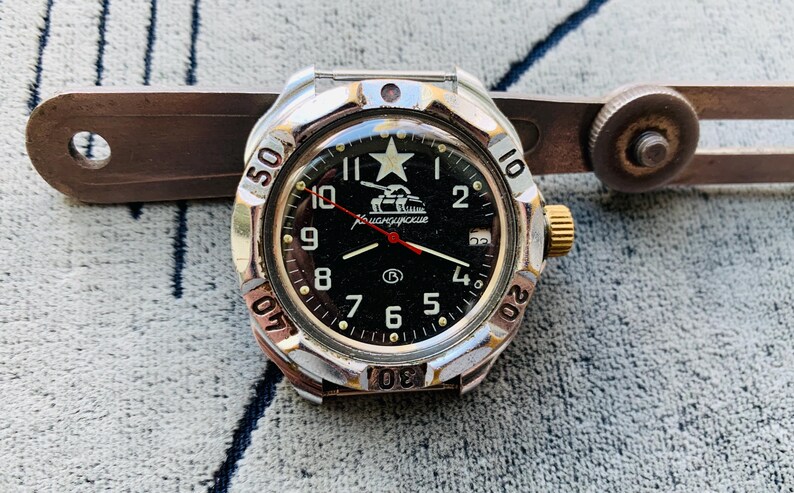 May include: A silver and gold-toned wristwatch with a black face, featuring white numerals and a star emblem. The watch has a red second hand and is set against a textured background. A metal tool is partially visible.