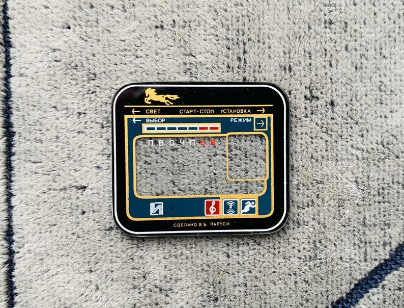 May include: A black and gold rectangular control panel with a horse silhouette and Cyrillic text. The panel has a blue and gold border, with icons and text in white and red. The panel is on a gray, textured surface.