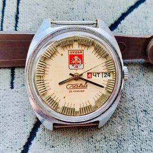 Könnte beinhalten: Eine silberfarbene Vintage-Armbanduhr mit cremefarbenem Zifferblatt. Die Uhr hat schwarze Zeiger, Stundenmarkierungen und kyrillischen Text. Sie ist an einem braunen Metallwerkzeug befestigt. Das Zifferblatt zeigt das Datum an.