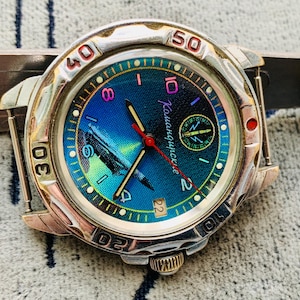 Op de afbeelding: Een zilverkleurig polshorloge met een blauw en groene wijzerplaat, met de tekst "Камандирские". Het horloge heeft een rode secondewijzer en een datumweergave. Het horloge ligt op een getextureerd oppervlak.