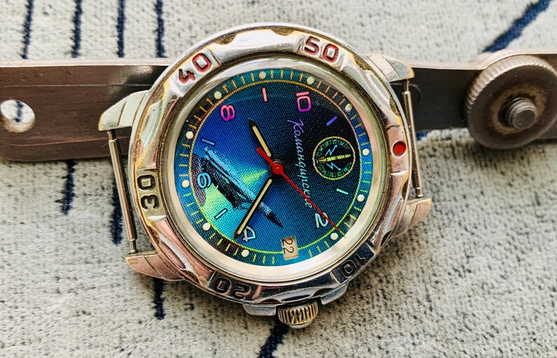 Op de afbeelding: Een zilverkleurig polshorloge met een blauw en groene wijzerplaat, met de tekst "Камандирские". Het horloge heeft een rode secondewijzer en een datumweergave. Het horloge ligt op een getextureerd oppervlak.