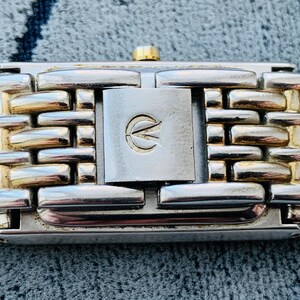 May include: Close-up of a watch band with a silver and gold-tone metal link design. The clasp is silver with a gold-tone accent and a logo. The watch band is on a textured gray surface.