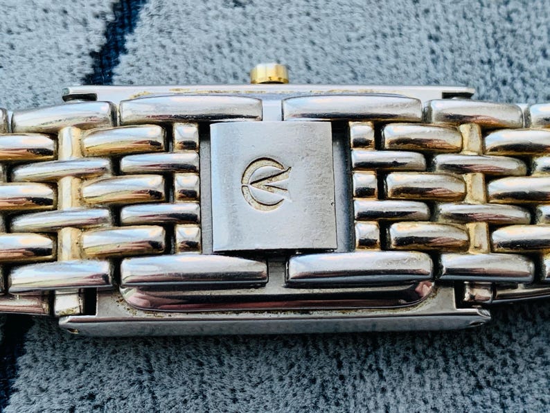 May include: Close-up of a watch band with a silver and gold-tone metal link design. The clasp is silver with a gold-tone accent and a logo. The watch band is on a textured gray surface.
