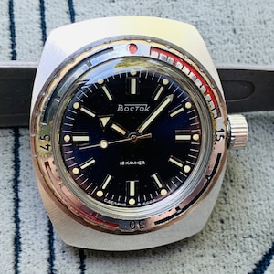 Peut inclure: Une montre-bracelet argentée avec un cadran noir et une lunette rouge et argentée. Le cadran de la montre comporte des marques blanches et le texte "Восток 18 камней Сделано в СССР" en écriture cyrillique.