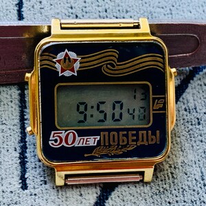 May include: A gold digital watch with a black face showing the time 9:50:48. The watch face has a red star emblem and the text "50 лет Победы" in gold. The watch is attached to a brown leather strap.