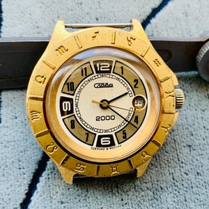 May include: A vintage gold-toned wristwatch with a cream-colored face. The watch features black hands, Arabic numerals, and a date display. The bezel is adorned with zodiac symbols. The watch reads "Слава" and "2000".