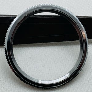 Op de afbeelding: Een zilveren horloge bezel met een gladde, gepolijste afwerking. De bezel heeft een reeks kleine, gelijkmatig verdeelde tanden rond de buitenrand.