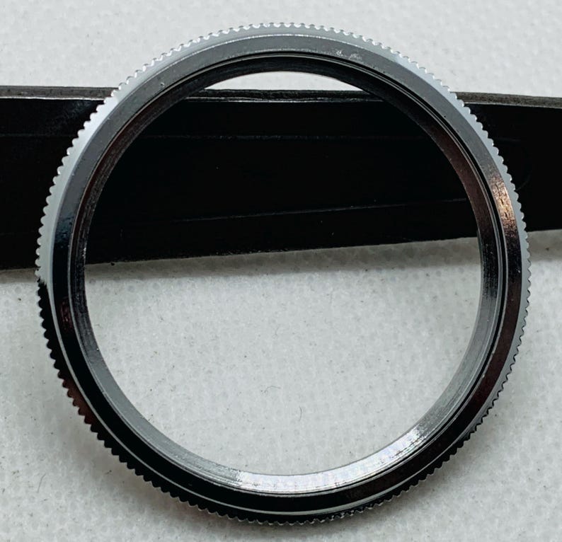 Op de afbeelding: Een zilveren horloge bezel met een gladde, gepolijste afwerking. De bezel heeft een reeks kleine, gelijkmatig verdeelde tanden rond de buitenrand.