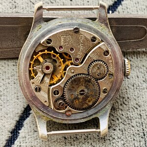 Puede incluir: Primer plano del mecanismo de un reloj vintage. El interior del reloj muestra intrincados engranajes, resortes y joyas en tonos dorados y plateados. La caja plateada del reloj tiene un aspecto desgastado, y la parte trasera est&aacute; abierta, revelando el mecanismo interno.