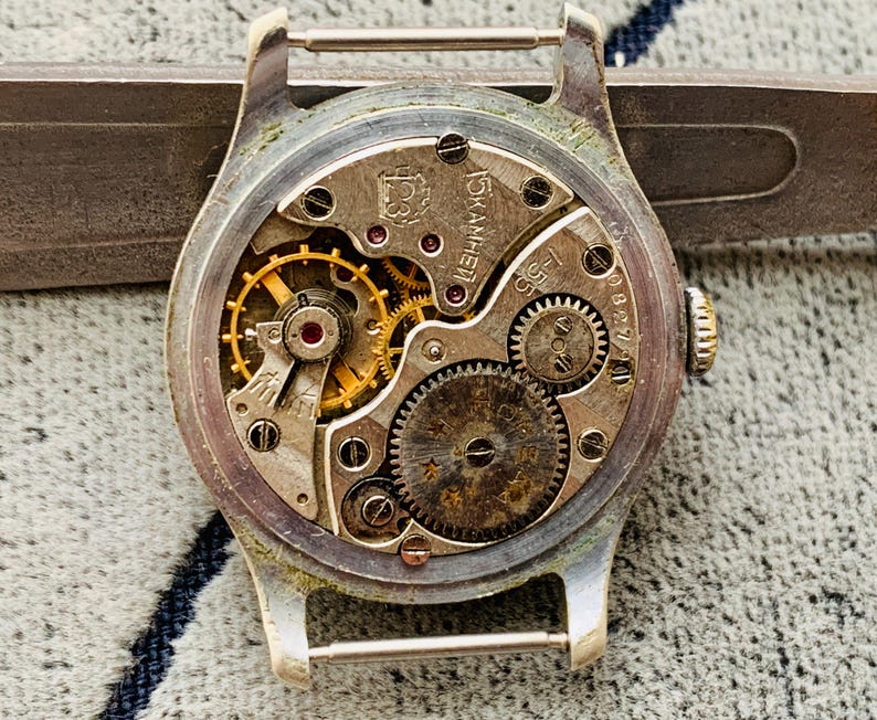 Puede incluir: Primer plano del mecanismo de un reloj vintage. El interior del reloj muestra intrincados engranajes, resortes y joyas en tonos dorados y plateados. La caja plateada del reloj tiene un aspecto desgastado, y la parte trasera est&aacute; abierta, revelando el mecanismo interno.