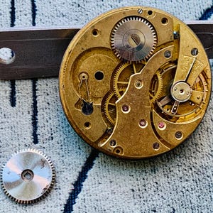 Puede incluir: Primer plano de un movimiento de reloj vintage con intrincados engranajes y componentes en tonos dorados y plateados. Una rueda dentada separada est&aacute; cerca. Una herramienta de metal est&aacute; colocada encima del movimiento del reloj.