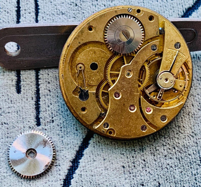 Puede incluir: Primer plano de un movimiento de reloj vintage con intrincados engranajes y componentes en tonos dorados y plateados. Una rueda dentada separada est&aacute; cerca. Una herramienta de metal est&aacute; colocada encima del movimiento del reloj.