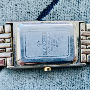 May include: A rectangular Perucci watch with a silver-tone metal band. The watch face is silver with a rectangular shape. The back of the watch has the text "STAINLESS STEEL BACK WATER PROOF JAPAN MOVT 23-1540".