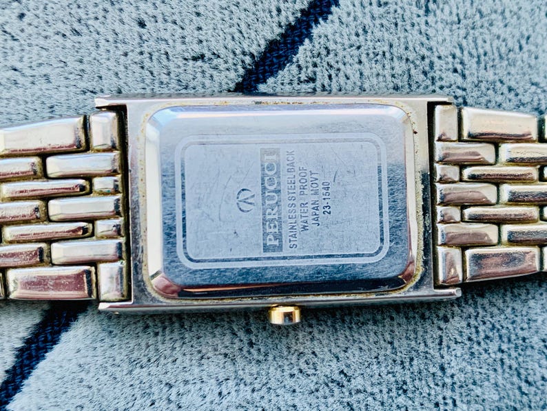 May include: A rectangular Perucci watch with a silver-tone metal band. The watch face is silver with a rectangular shape. The back of the watch has the text "STAINLESS STEEL BACK WATER PROOF JAPAN MOVT 23-1540".