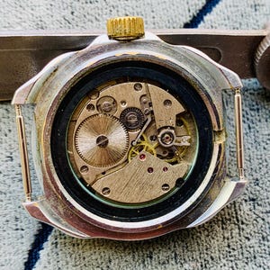 May include: Close-up of a vintage watch mechanism. The watch's back is open, revealing intricate gears, springs, and jewels. The watch case is silver-toned with a gold crown. The watch is being held by a silver tool.