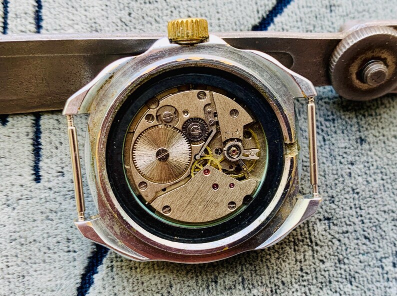 May include: Close-up of a vintage watch mechanism. The watch's back is open, revealing intricate gears, springs, and jewels. The watch case is silver-toned with a gold crown. The watch is being held by a silver tool.