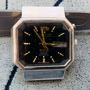 May include: A silver-toned Orient Crystal 21 Jewels watch with a black face. The watch has gold-colored hands and markers, and a day-date display. The watch is rectangular with an octagonal bezel.