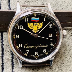 May include: A black wristwatch with a silver band and a black face. The face has white numbers and hands, a small date window, and a gold eagle emblem with a Russian flag above it. The text "Спецназовские" is printed in gold on the face.