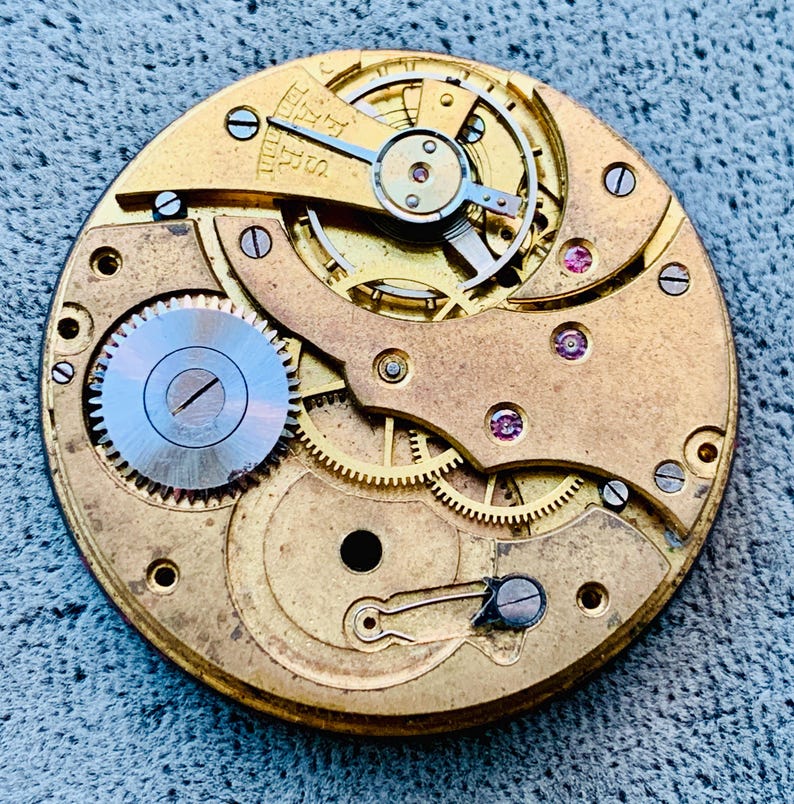 Puede incluir: Primer plano de un movimiento de reloj antiguo, que muestra intrincados engranajes, resortes y joyas. Los componentes de color lat&oacute;n est&aacute;n dispuestos en un patr&oacute;n circular, con un engranaje plateado en el centro. Un ejemplo de ingenier&iacute;a de precisi&oacute;n.
