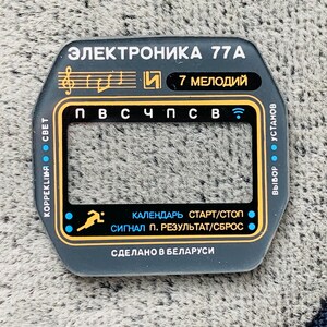 May include: A dark gray watch face with gold and white text. The text includes "ЭЛЕКТРОНИКА 77A", "7 МЕЛОДИЙ", and Cyrillic characters. It features a rectangular display area and various function labels. Made in Belarus.