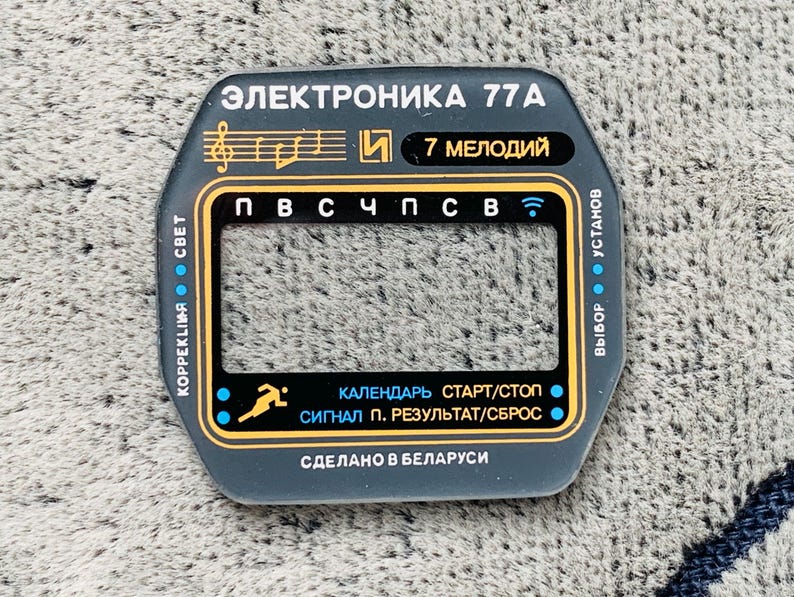 May include: A dark gray watch face with gold and white text. The text includes "ЭЛЕКТРОНИКА 77A", "7 МЕЛОДИЙ", and Cyrillic characters. It features a rectangular display area and various function labels. Made in Belarus.