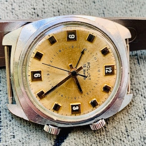 Puede incluir: Un reloj de pulsera vintage con esfera dorada, manecillas negras e indicadores horarios rectangulares negros. La esfera del reloj muestra los n&uacute;meros 3, 6, 9 y 12. El reloj tiene una caja plateada y una correa de cuero marr&oacute;n.