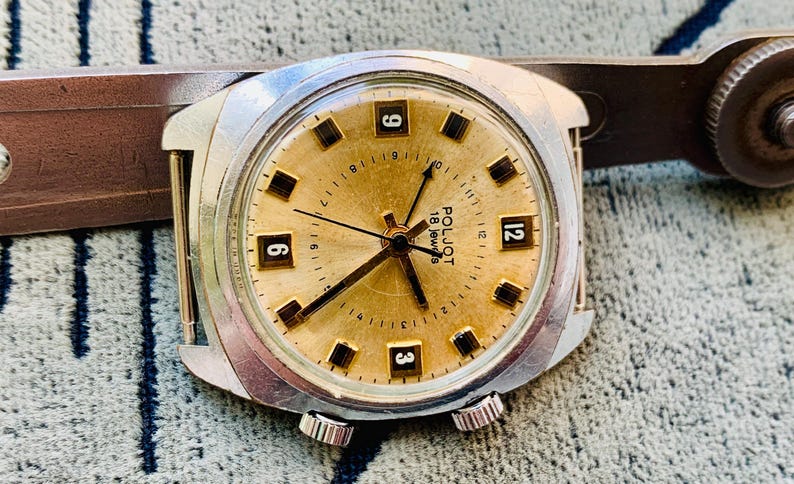 Puede incluir: Un reloj de pulsera vintage con esfera dorada, manecillas negras e indicadores horarios rectangulares negros. La esfera del reloj muestra los n&uacute;meros 3, 6, 9 y 12. El reloj tiene una caja plateada y una correa de cuero marr&oacute;n.