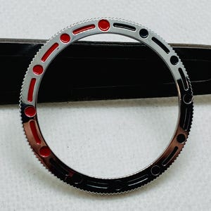 Op de afbeelding: Een zilveren horloge bezel met rode en zwarte markeringen. De bezel is ontworpen voor een horloge met een draaibare buitenring.
