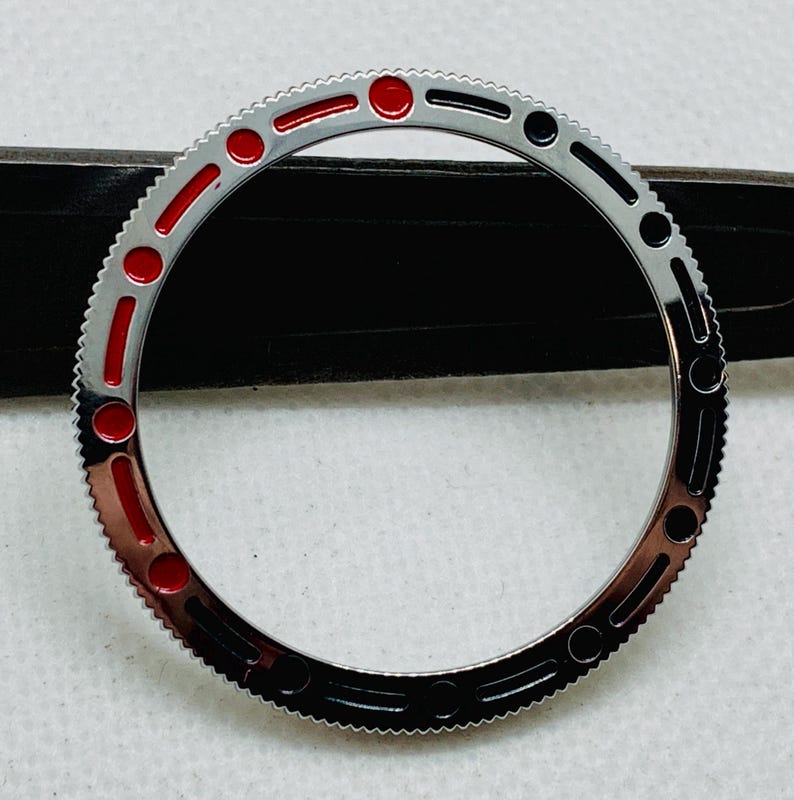 Op de afbeelding: Een zilveren horloge bezel met rode en zwarte markeringen. De bezel is ontworpen voor een horloge met een draaibare buitenring.