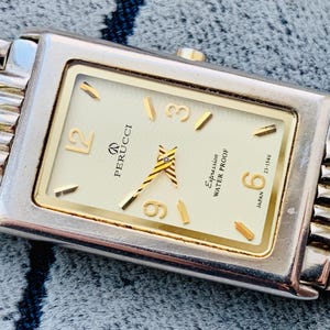 May include: A rectangular Perucci watch with a silver and gold band. The watch face is cream-colored with gold accents, including the hands and hour markers. The watch is labeled "Expression Water Proof" and "Japan".