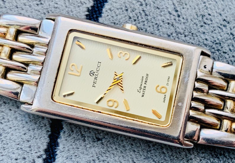 May include: A rectangular Perucci watch with a silver and gold band. The watch face is cream-colored with gold accents, including the hands and hour markers. The watch is labeled "Expression Water Proof" and "Japan".