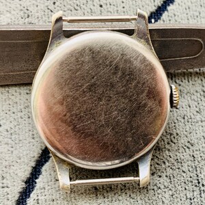 Puede incluir: Primer plano de la parte trasera de la caja de un reloj de pulsera vintage. El reloj tiene una caja redonda de metal cepillado con una peque&ntilde;a corona. La caja est&aacute; sujeta a un soporte de correa de metal. El reloj est&aacute; sobre una superficie texturizada.