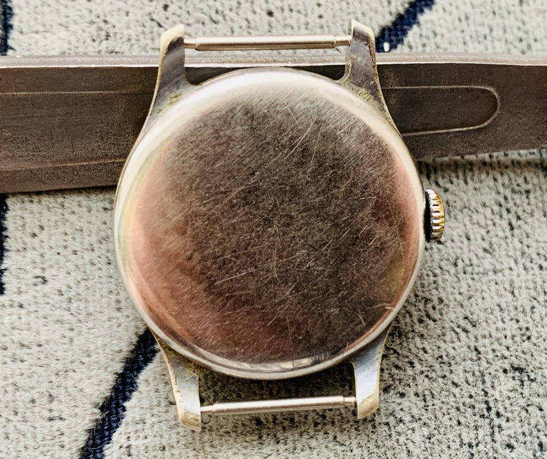 Puede incluir: Primer plano de la parte trasera de la caja de un reloj de pulsera vintage. El reloj tiene una caja redonda de metal cepillado con una peque&ntilde;a corona. La caja est&aacute; sujeta a un soporte de correa de metal. El reloj est&aacute; sobre una superficie texturizada.