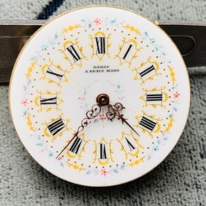 K&ouml;nnte beinhalten: Ein Vintage-Zifferblatt mit r&ouml;mischen Ziffern und zarten floralen Akzenten. Das wei&szlig;e Zifferblatt hat schwarze Ziffern, goldfarbene Verzierungen und den Text "HARDY A PETIT MARS". Die Zeiger sind verziert, und das Design umfasst rosa und blaue Blumenmuster.