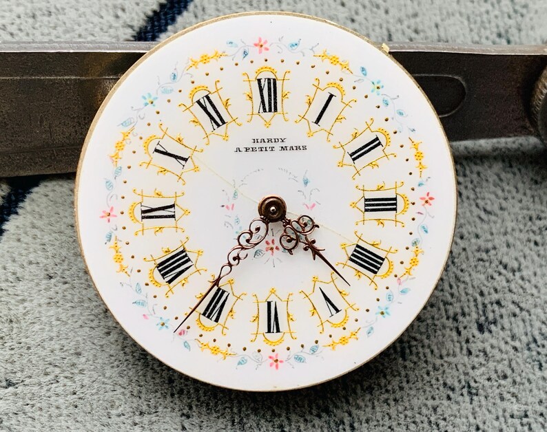K&ouml;nnte beinhalten: Ein Vintage-Zifferblatt mit r&ouml;mischen Ziffern und zarten floralen Akzenten. Das wei&szlig;e Zifferblatt hat schwarze Ziffern, goldfarbene Verzierungen und den Text "HARDY A PETIT MARS". Die Zeiger sind verziert, und das Design umfasst rosa und blaue Blumenmuster.