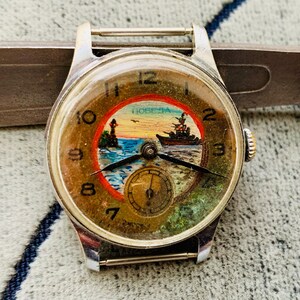 Puede incluir: Reloj de pulsera vintage con caja plateada y esfera redonda. La esfera del reloj presenta una escena n&aacute;utica con un barco, un faro y la palabra "POBEDA". El reloj tiene agujas negras y una subesfera en la parte inferior.